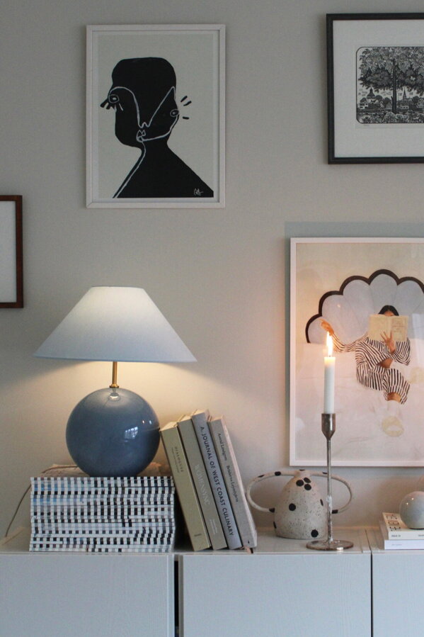 Modrá stolová lampa na bielej komode s knihami a rôznymi dekoráciami pod stenou s rôznymi obrázkami.