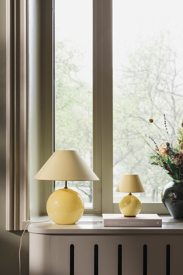 Menšia a väčšia svetložltá stolová lampa na stole vedľa čiernej vázy s kyticou kvetou, pred oknom.