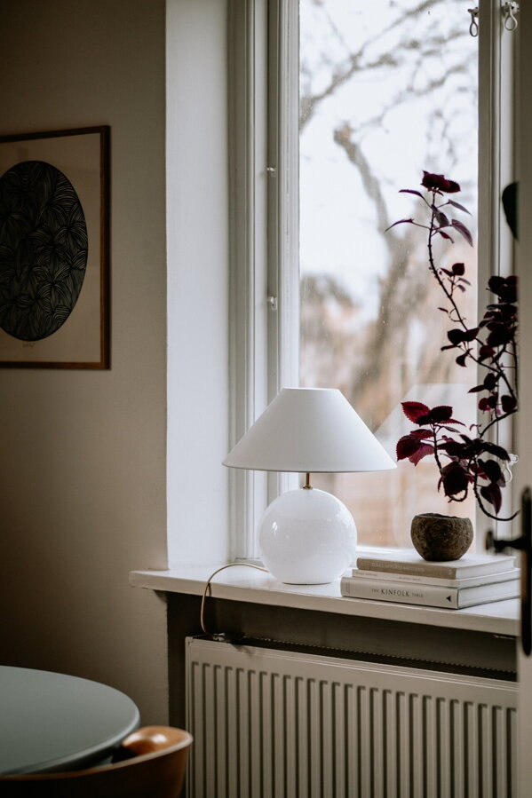 Biela stolová lampa s guľatou sklenenou základňou položená na parapete okna vedľa kôpky kníh a izbovej rastliny.