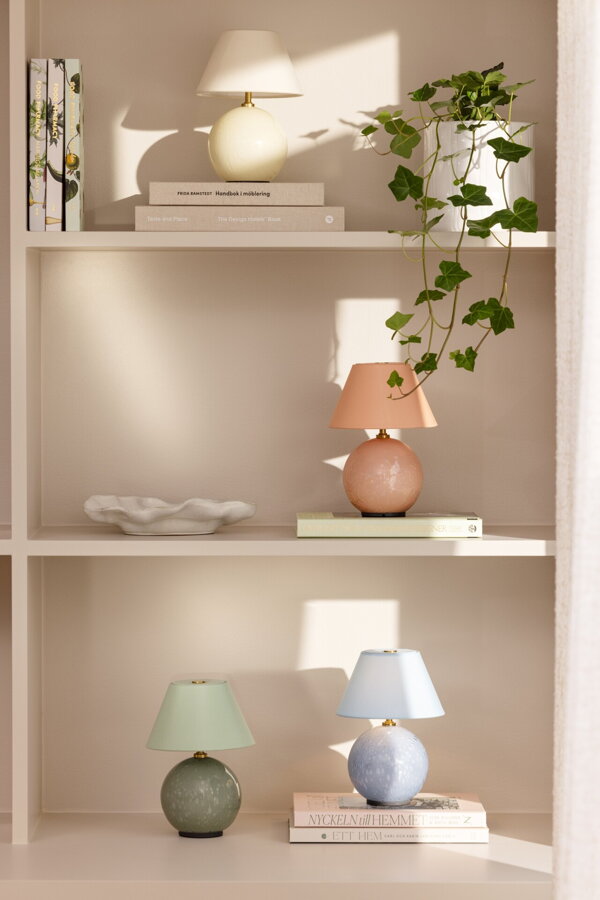 Svetloružová svietiaca prenosná stolová lampa Iris položená bielej skrinke pod policami.