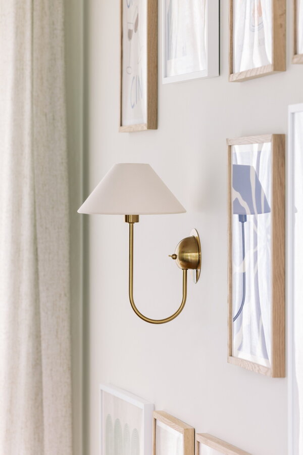 Nástenná lampa Iris s kovovou konštrukciou v prevedení brúsenej mosadze a s krémovým bavlneným tienidlom, na stene medzi viacerými obrazmi.