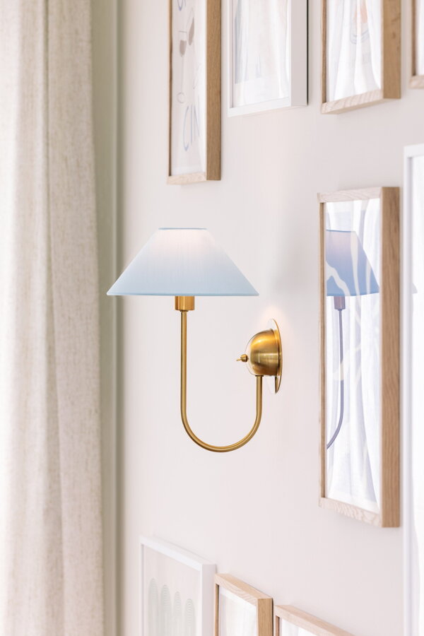 Svietiaca nástenná lampa Iris s kovovou konštrukciou v prevedení brúsenej mosadze a so svetlomodrým bavlneným tienidlom, na stene medzi viacerými obrazmi.