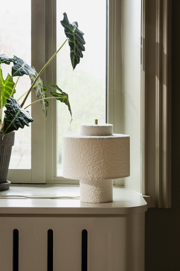 Stolová lampa Noah s bielym tienidlom z tkaniny bouclé a prvkami v prevedení pravej mosadze na parapete okna.