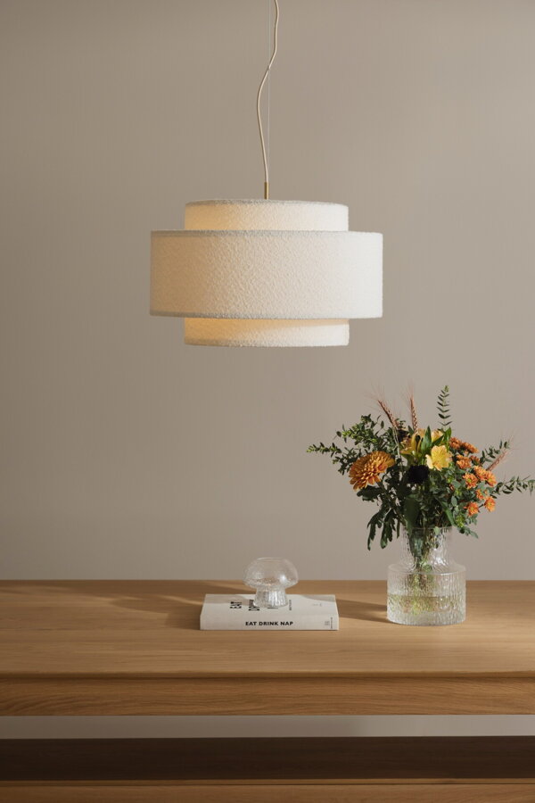 Svietiaca závesná lampa Noah s bielym tienidlom z tkaniny bouclé a prvkami v prevedení pravej mosadze nad dreveným stolom, na ktorom je váza s kvetmi.