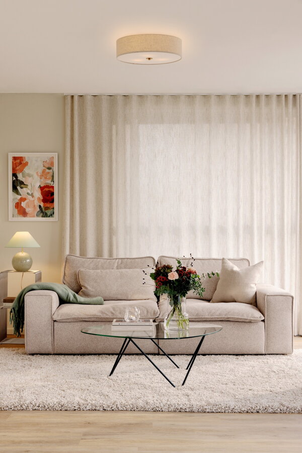 Stropná lampa Noah s béžovým tienidlom z kvalitného ľanu a prvkami v prevedení pravej mosadze v obývačke nad béžovou sedačkou.