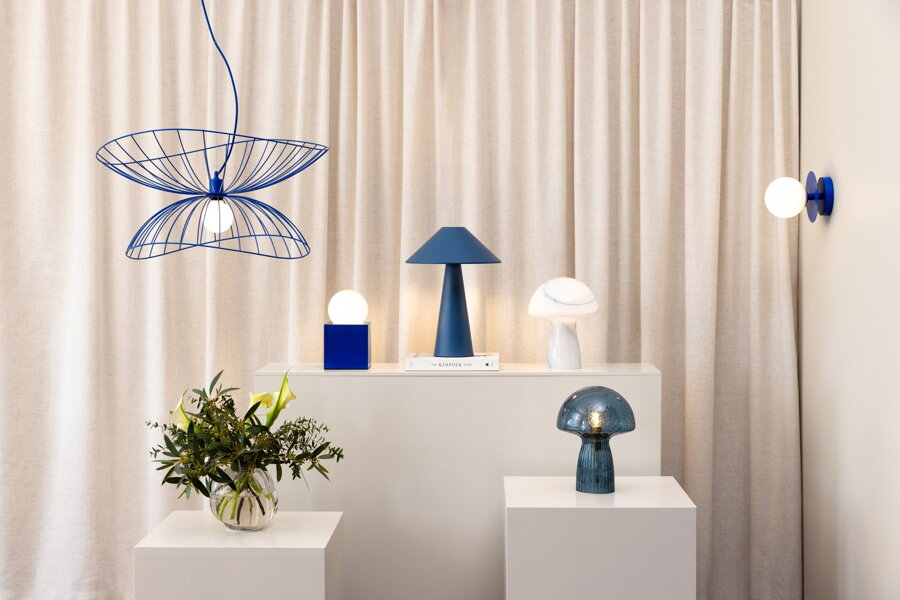 Viacero modrých svietidiel rôzneho druhu od závesných, stolových a nástenných, ktoré su rozložené na bielych podstavcoch.