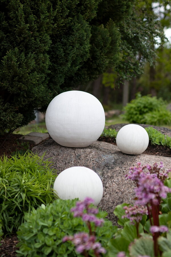 Tri biele guľaté vonkajšie lampy v záhrade medzi záhonmi kvetov.