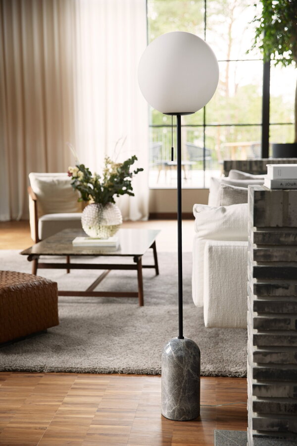 Stojanová lampa Torrano so sivou mramorovou základňou a s jemným tienidlom z bieleho opálového skla na podlahe vedľa bielej pohovky v obývačke.