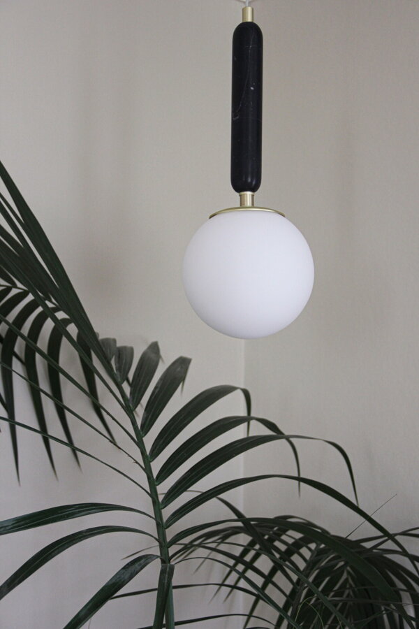 Závesná lampa Torrano s masívnou čiernou mramorovou základňou a s jemným tienidlom z bieleho opálového skla nad izbovou rastlinou.