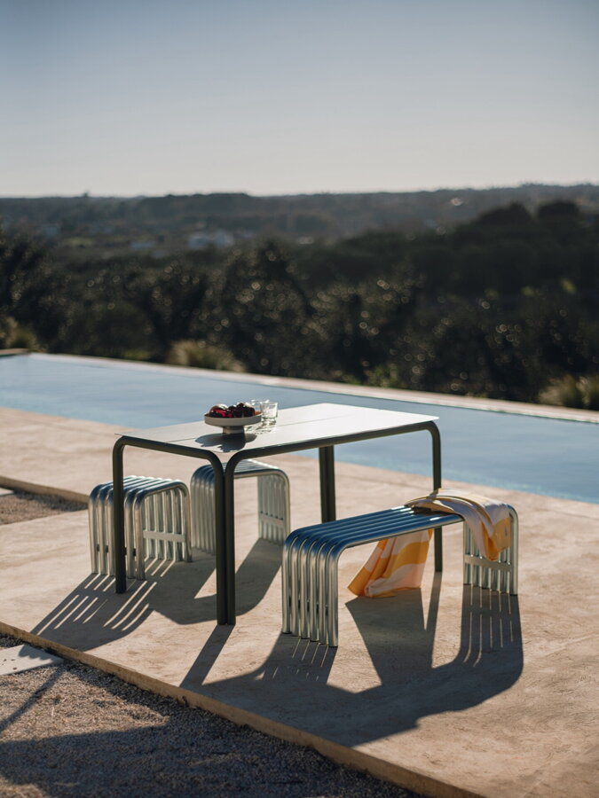 Kovová vonkajšia lavica v striebornnej farbe vedľa kovového stolíka pri bazéne.