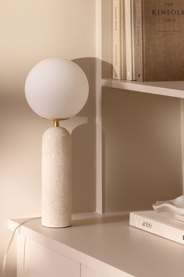 Stolová lampa Torrano s masívnou travertínovou základňou a s jemným tienidlom z bieleho opálového skla, na bielej skrinke.