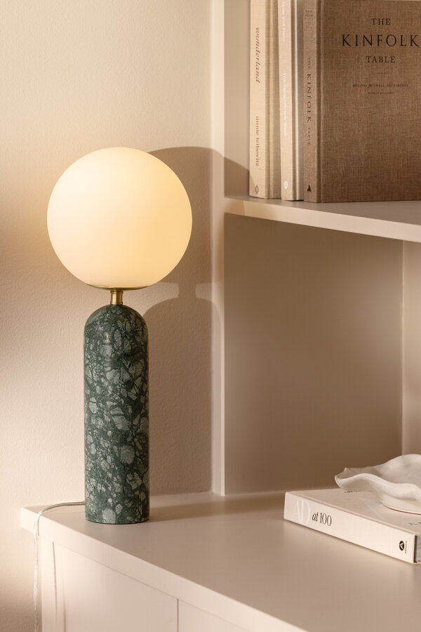 Svietiaca stolová lampa Torrano s tmavozelenou mramorovou základňou a s jemným tienidlom z bieleho opálového skla, na bielej skrinke.
