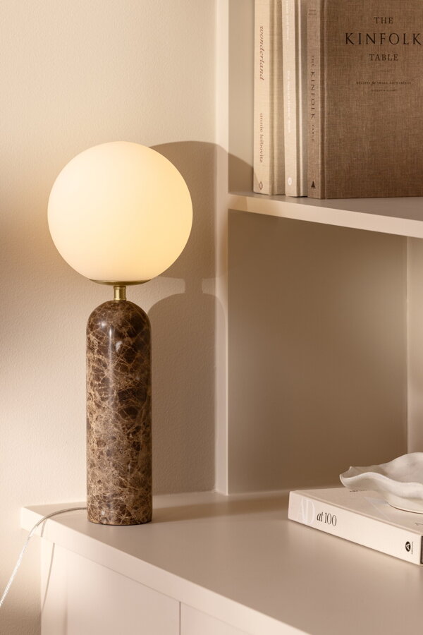 Svietiaca stolová lampa Torrano s hnedou mramorovou základňou a s jemným tienidlom z bieleho opálového skla, na bielej skrinke.