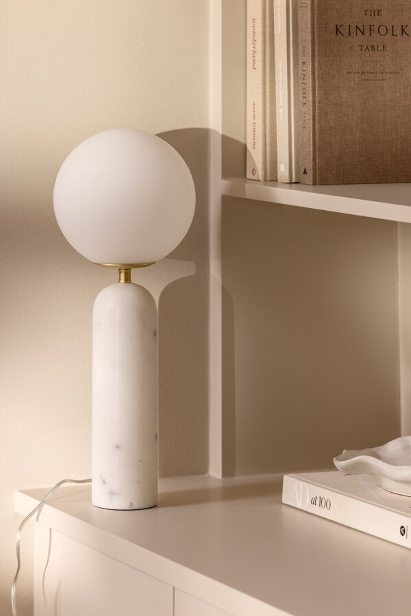 Stolová lampa Torrano s bielou mramorovou základňou a s jemným tienidlom z bieleho opálového skla, na bielej skrinke.