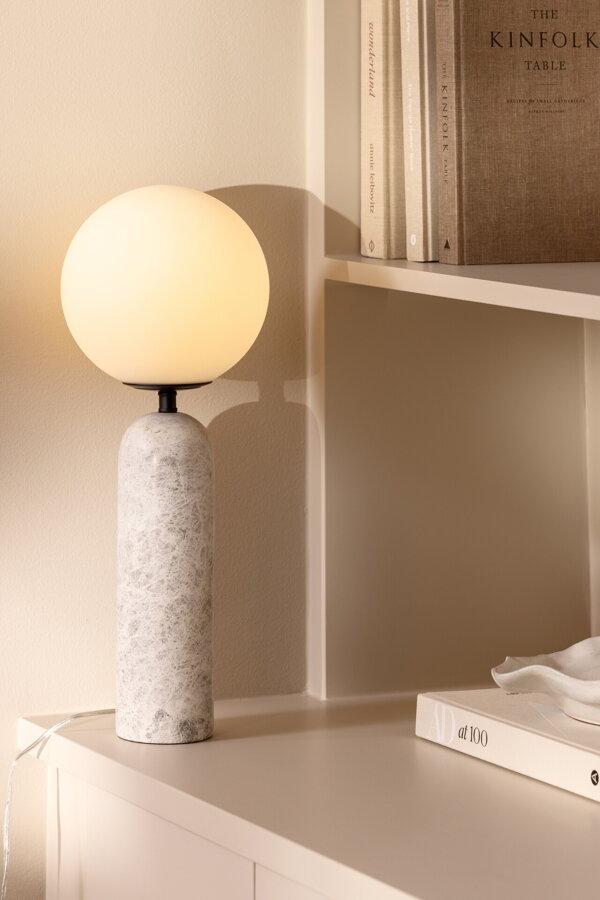Svietiaca stolová lampa Torrano so sivou mramorovou základňou a s jemným tienidlom z bieleho opálového skla, na bielej skrinke.
