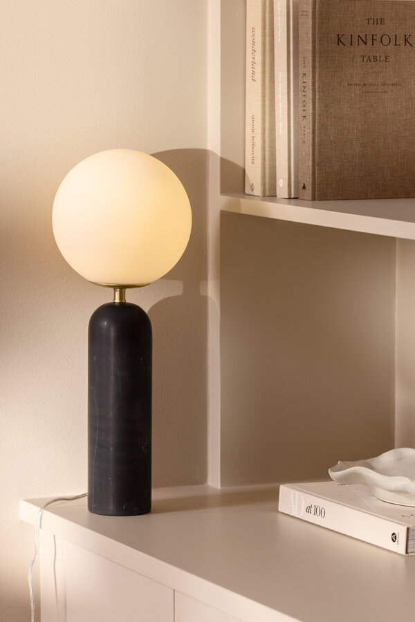 Svietiaca stolová lampa Torrano s čiernou mramorovou základňou a s jemným tienidlom z bieleho opálového skla, na bielej skrinke.