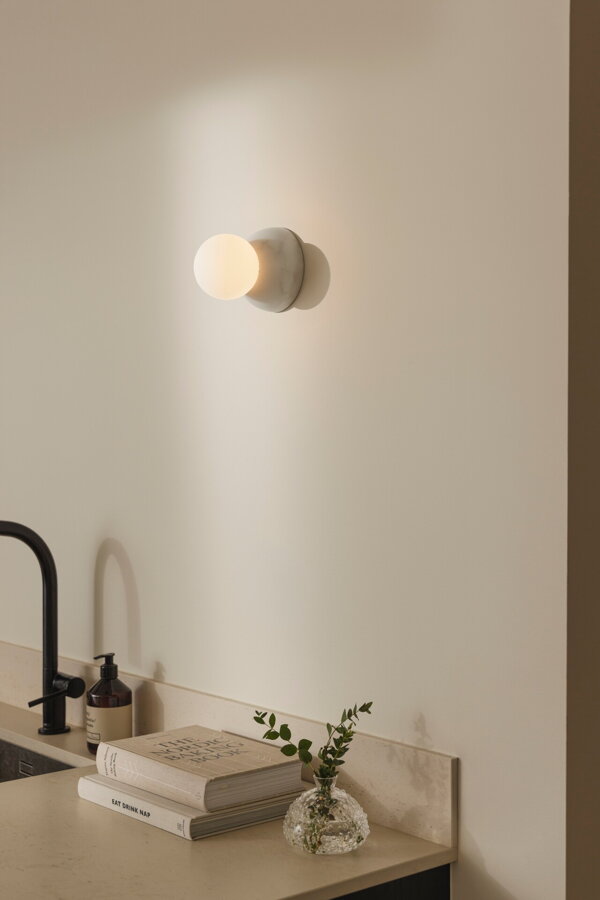 Nástenná lampa Torrano s maso bielou mramorovou základňou a s jemným tienidlom z bieleho opálového skla v kuchyni nad umývadlom.