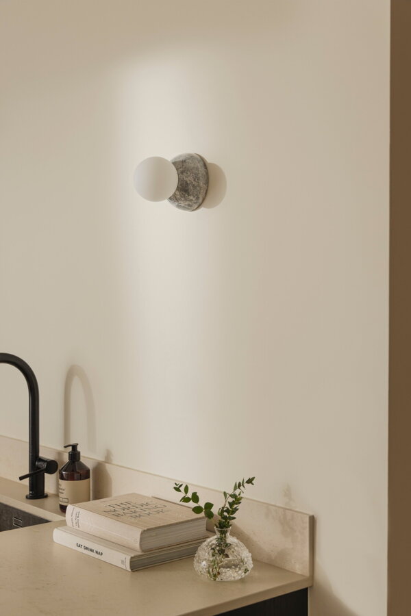 Nástenná lampa Torrano s masívnou sivou mramorovou základňou a s jemným tienidlom z bieleho opálového skla na stene v kuchyni nad umývadlom.