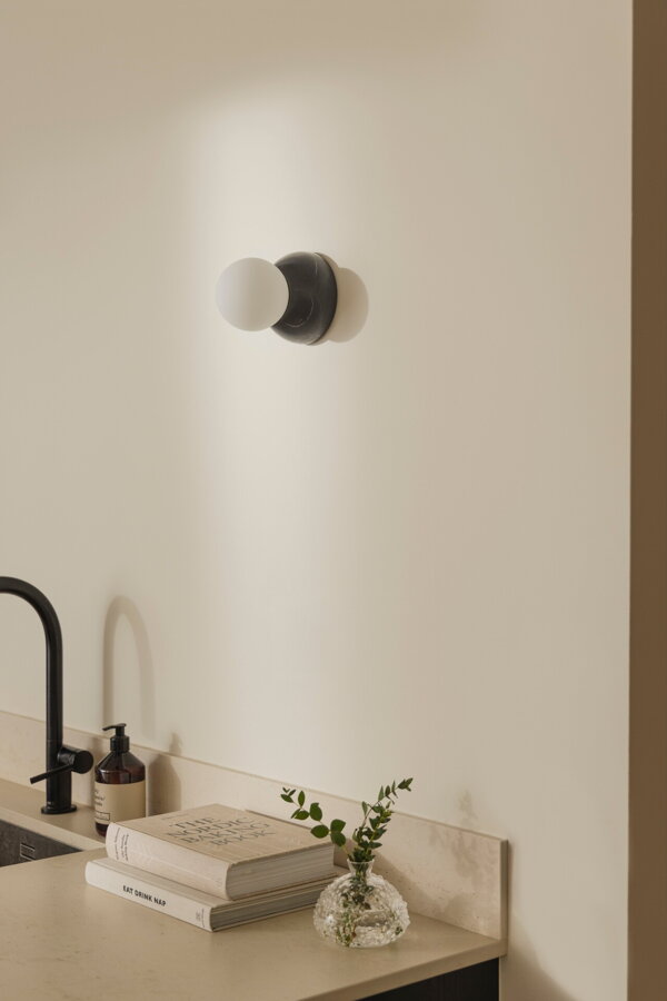 Nástenná lampa Torrano s masívnou čiernou mramorovou základňou a s jemným tienidlom z bieleho opálového skla na stene v kuchyni nad umývadlom.