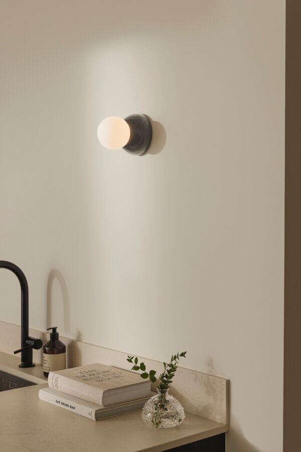 Nástenná lampa Torrano s masívnou čiernou mramorovou základňou a s jemným tienidlom z bieleho opálového skla na stene v kuchyni nad umývadlom.