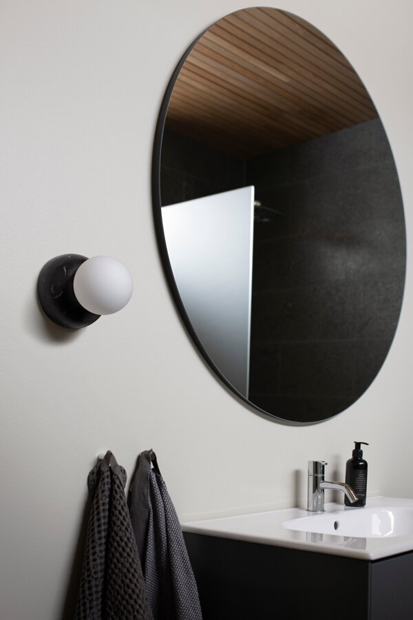 Nástenná lampa Torrano s masívnou čiernou mramorovou základňou a s jemným tienidlom z bieleho opálového skla na stene v kúpeľni vedľa umývadla a zrkadla.