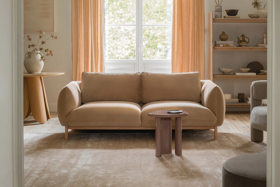 2-miestna pohovka pieskovej farby v obývačke s hnedým lesklým príručným stolíkom, v pozadí s vysokou policou s dekoráciami.