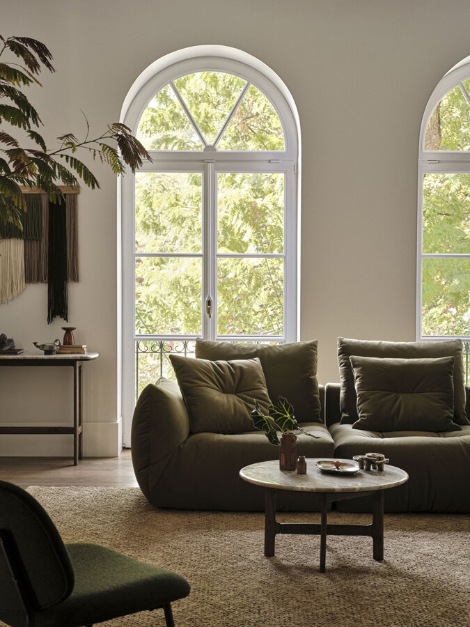 Svetlozelená rohová modulárna sedačka v obývačke s vysokými oblúkovými oknami a vysokou policovou stenou.