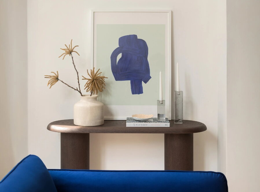 Tmavohnedý oválny konzolový stolík Bufi s vázou s kvetmi pri stene, na ktorej je zavesený obraz.