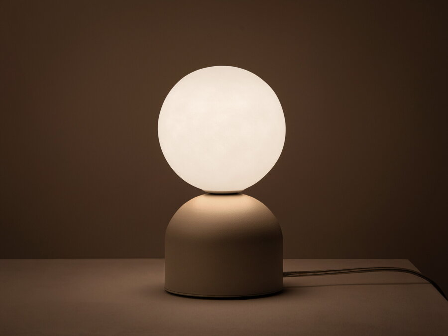 V tme rozsvietená stolová lampa Ely s matnou béžovou kovovou základňou a matným bielym skleneným tienidlom v tvare gule.