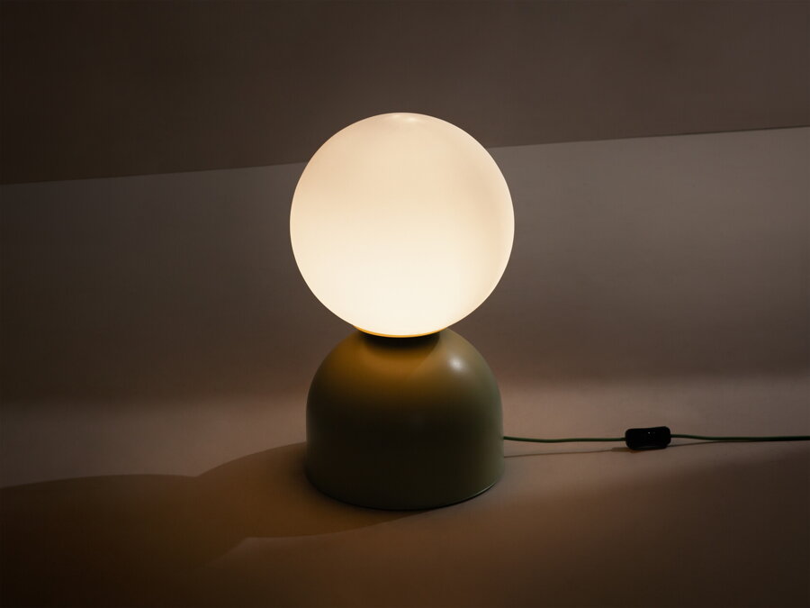 V tme rozsvietená stolová lampa Ely s matnou svetlozelenou kovovou základňou a matným bielym skleneným tienidlom v tvare gule.