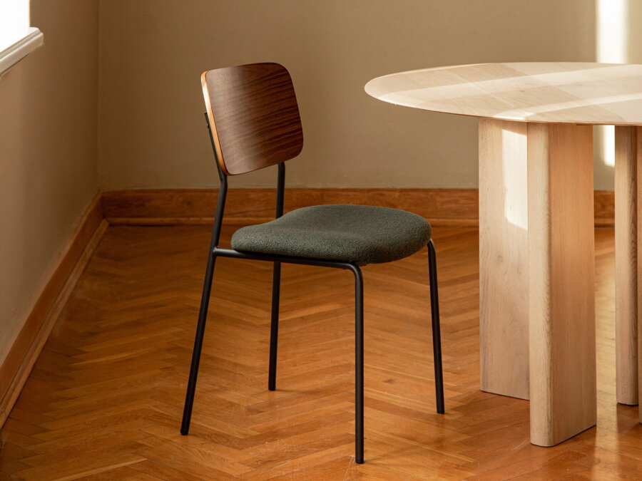 Khaki zelená čalúnená jedálenská stolička s dreveným operadlom a čiernymi kovovými nohami pri svetlom jedálenskom stole.