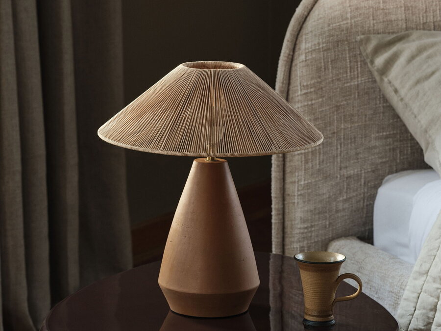 Stolová lampa s terakotovou základňou a tienidlom z viskózového povrazu na nočnom stolíku pri posteli..