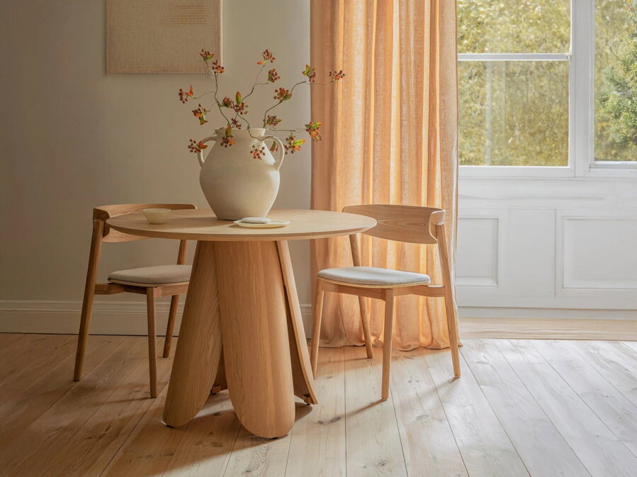 Dve prírodné jaseňové jedálenské stoličky s čalúneným béžovým sedákom za okrúhlym jaseňovým jedálenským stolom.