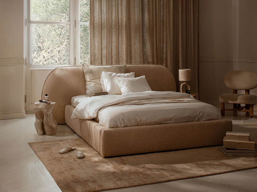 Béžová čalúnená manželská posteľ v spálni s veľkým francúzskym oknom, na béžovom koberci.