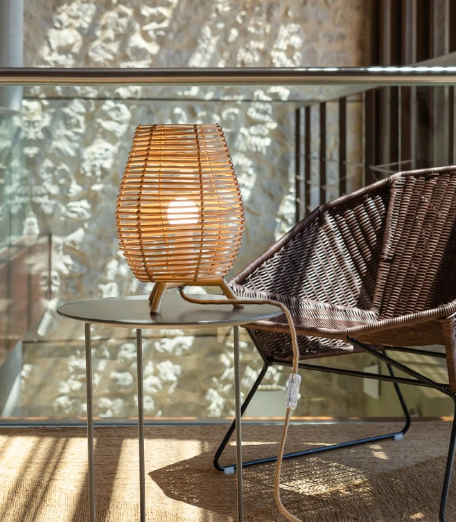 Stolová ratanová lampa na stole vedľa ratanoveho kresla v žiare slnečných lúčov