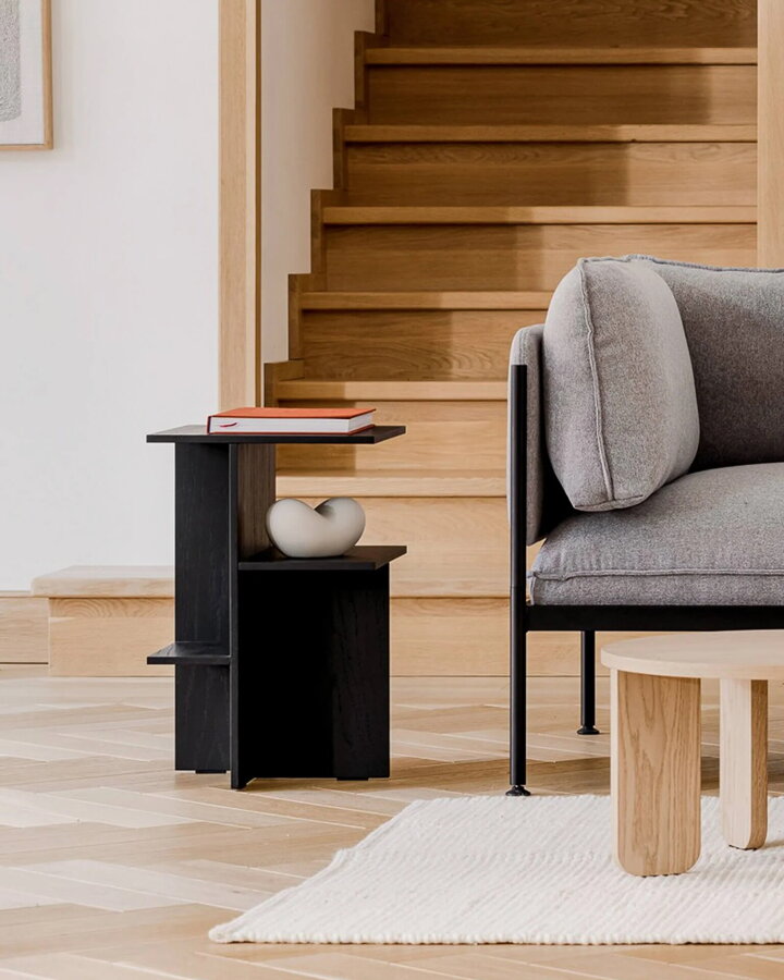 Čierny dubový nočný stolík Atik v obývačke pred dreveným schodiskom vedľa sivej pohovky a konferenčného stolíka.