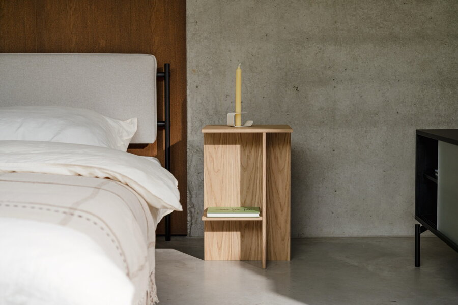 Dubový nočný stolík vedľa ustlanej postele, na ktorom je položený svietnik so žltou sviečkou.