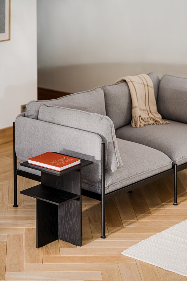 Elegantný nočný stolík Atik v čiernej farbe z matnej dubovej dyhy v obývačke vedľa sivej pohovky.