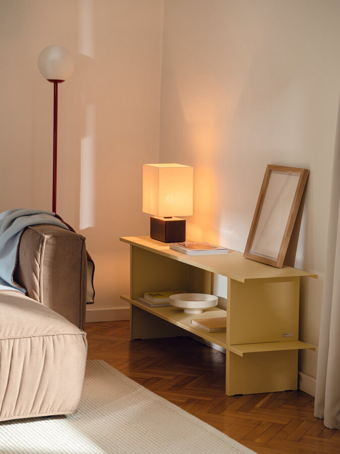 Nízka svetložltá polica v spálni, na polici je nočná lampa a rámik s fotkou.