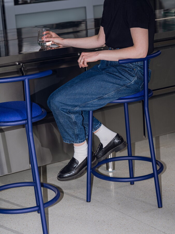Žena v džínsoch a čiernom tričku sediaca na modrej barovej stoličke s opierkou.