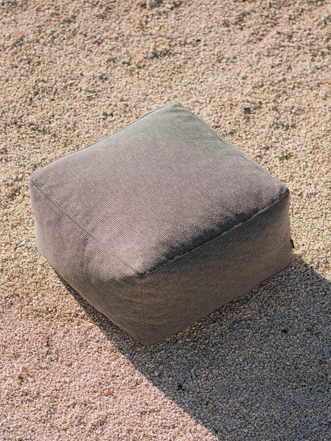 Moderný čalúnený puf Keno Dusty Beige v sivej farbe, so snímateľným poťahom, vhodný do exteriéru, položený na piesku.