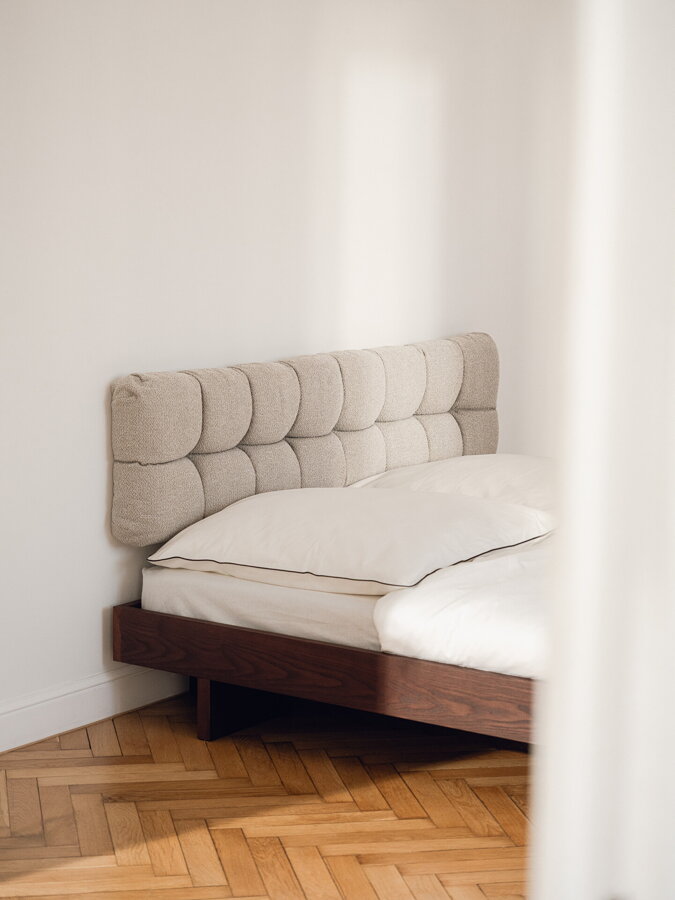 Tmavohnedá drevená manelská posteľ v spálni, na stene za posteľou je béžové čalúnené čelo na stene.