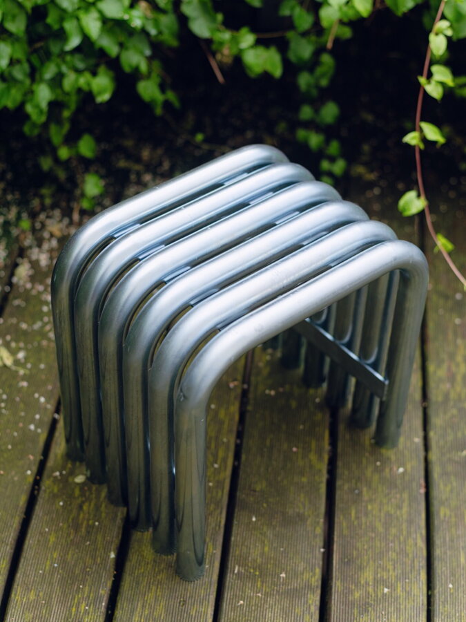 Stolík/stolička Nokk v striebornej farbe z galvanizovanej ocele v exteriéri pri zelenom kry na drevenej podlahe.