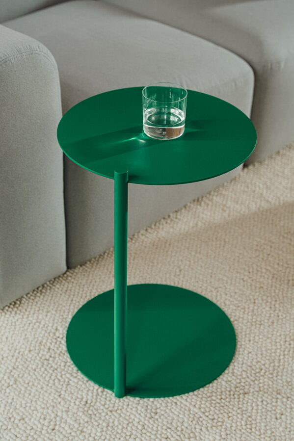 Oceľový príručný stolík Ande zelenej farby so skleneným pohárom vody vedľa svetlosivej pohovky.