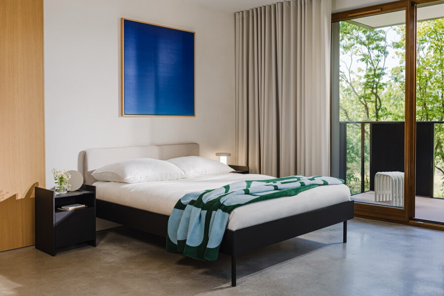 Čierna melamínová manželská posteľ s čalúneným pieskovým čelom v spálni na drevenej podlahe, nad posteľou visí veľký modrý abstraktný obraz.