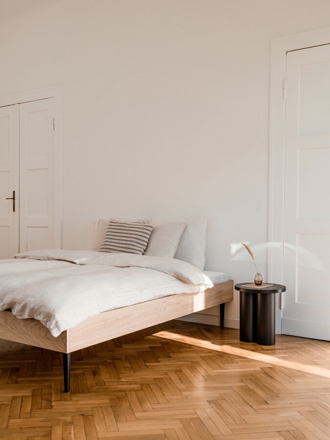 Dubová manželská posteľ v spálni na drevenej podlahe vedľa čierneho príručného stolíka.