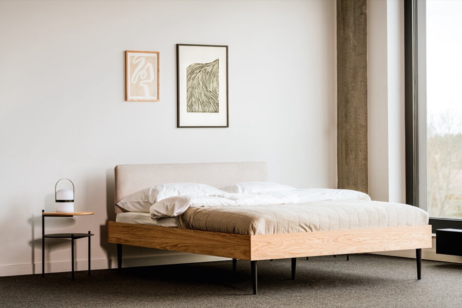 Dubová manželská posteľ s pieskovým čalúneným čelom vo svetlej spálni na sivej podlahe vedľa čierneho nočného stolíka.