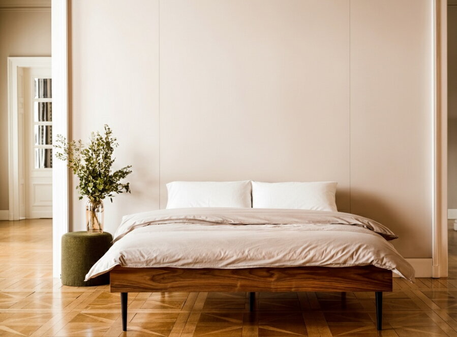 Orechová manželská posteľ vo svetlej spálni na drevenej podlahe vedľa zeleného príručného stolíka s vázou s kvetmi.