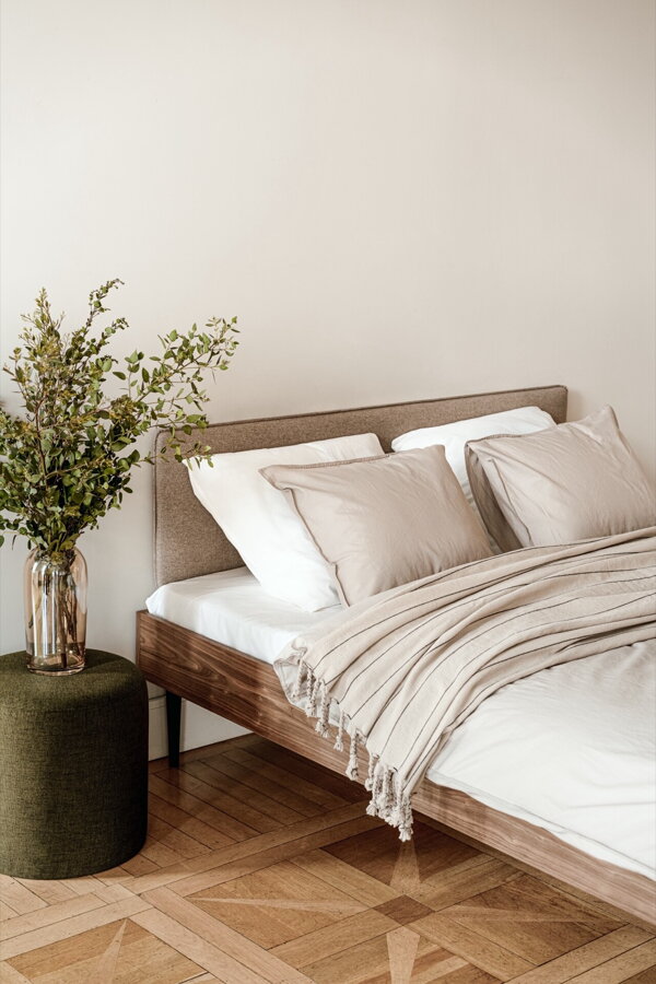 Orechová manželská posteľ s kávovo hnedým čalúneným čelom vo svetlej spálni na drevenej podlahe vedľa zeleného príručného stolíka s vázou s kvetmi.