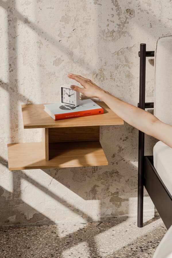 Minimalistický nočný stolík Tann s dvoma úrovňami z matnej dubovej dyhy prírodnej farby zavesený na stene vedľa postele, na stolíku je kniha a budík.